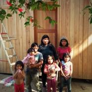Mulher e seis crianças do povo Guarani em frente a casa de madeira.