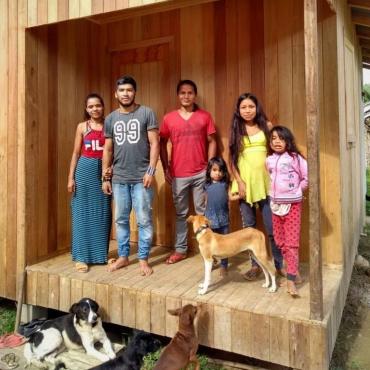 Duas mulheres, dois homens, duas meninas e alguns cachorros em frente a casa de madeira.