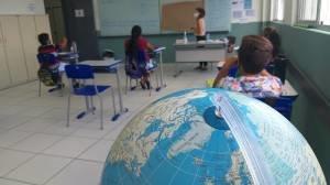 Foto da sala de aula, com um globo no primeiro plano e Karina conversando com os alunos no fundo.