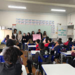 Grupo de alunas em frente da sala de aula segurando um cartaz rosa e falando para uma sala de aula cheia de alunos.