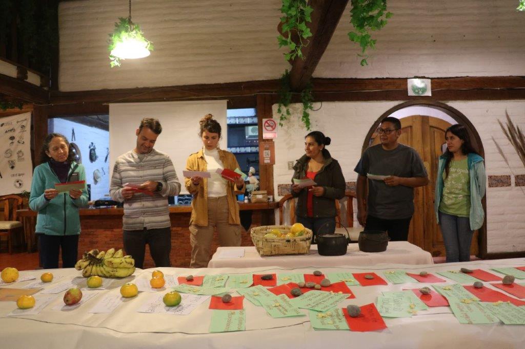 No primeiro plano uma mesa na qual há uma cartolina branca estendida e sob ela cartões verdes e vermelhos. Ao fundo, os técnicos quatro mulheres e dois homens de pé.