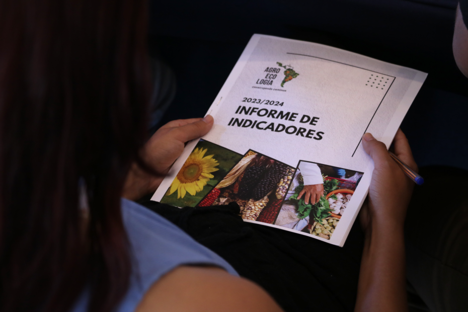 Mãos femininas seguram um relatório. Na capa se lê: "Relatório de Indicadores 2023 / 2024"